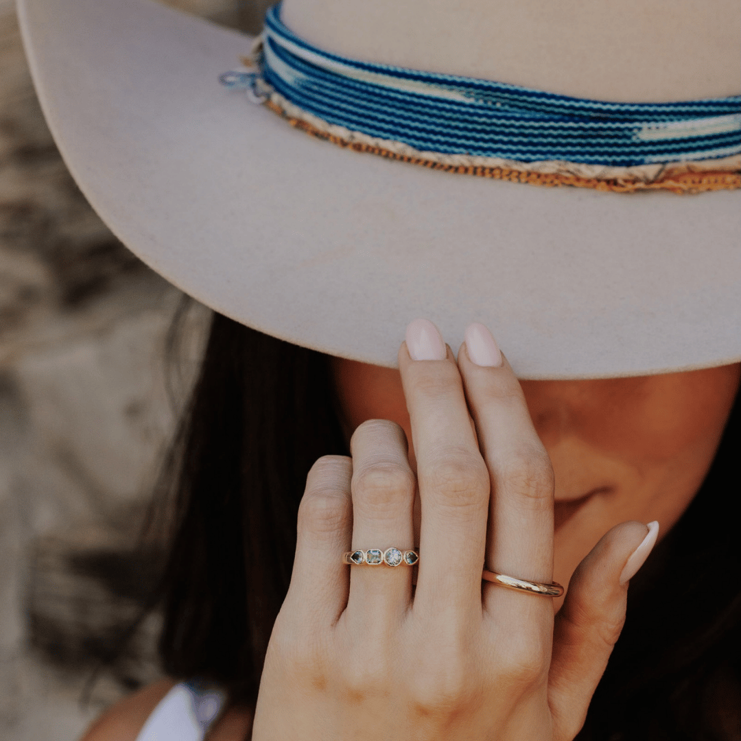 A 14k gold ring featuring four blue sapphire gemstones, displayed on the ring finger of a hand model tipping her hat. 
