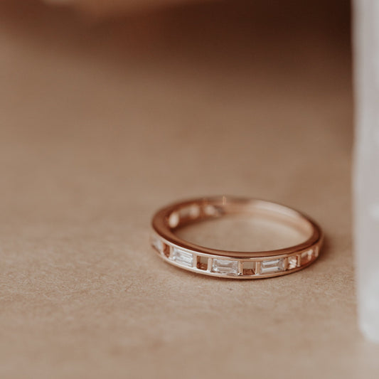 Rose gold wedding band with baguette diamond accents on a beige surface.