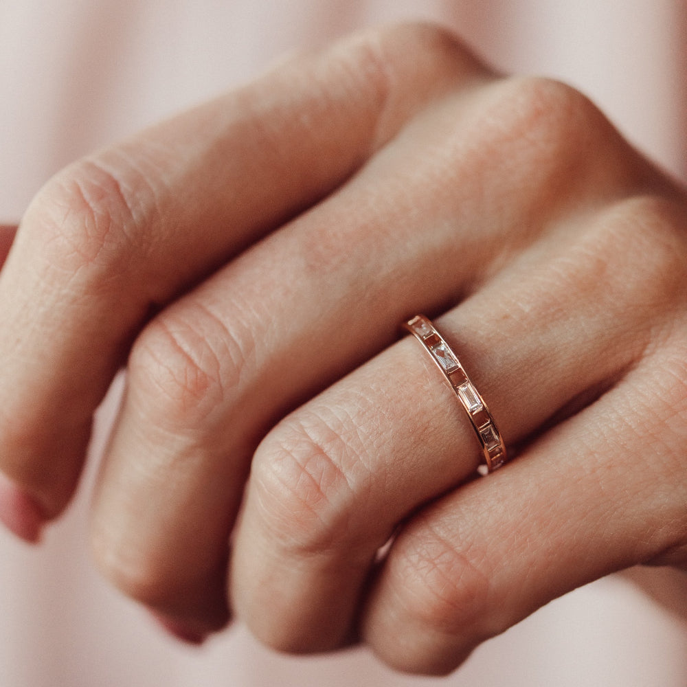 Rose gold Bridget Diamond wedding band on a person's finger with a soft pink background.