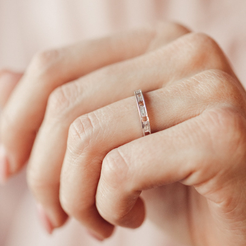 Close-up of a hand wearing a diamond wedding band on a soft pink background.
