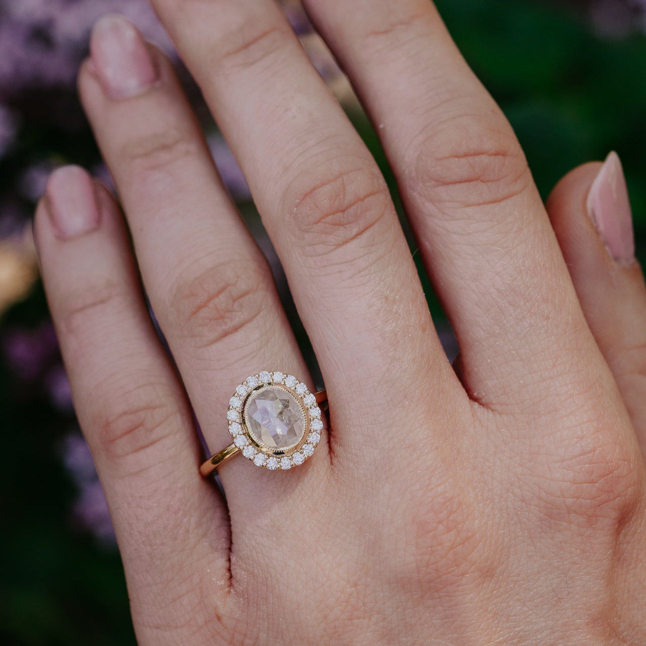 Hand wearing a ring with a clear oval rustic diamond halo ring against a blurred natural background.