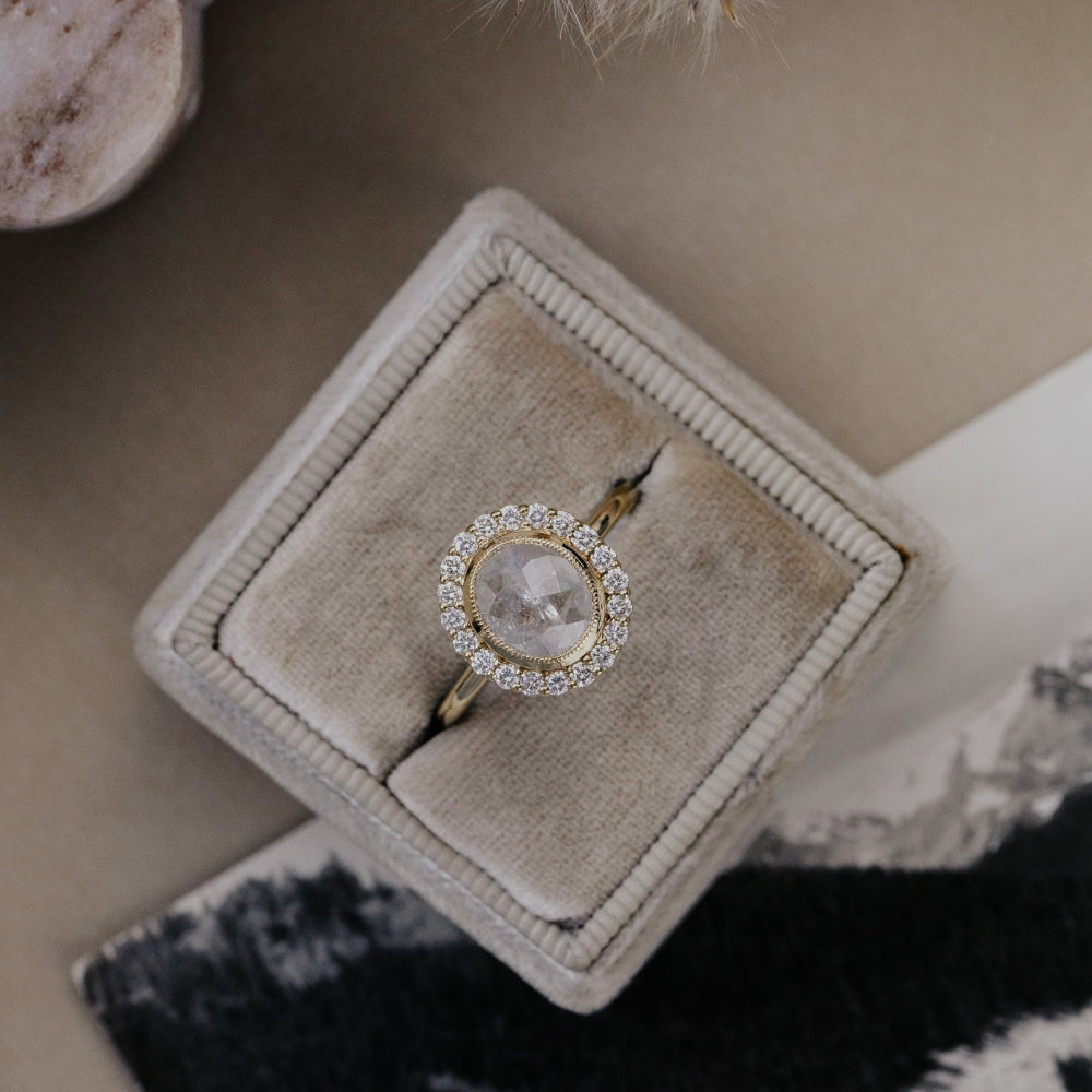 Gold ring with a large oval rustic diamond center stone and a diamond halo, placed on a beige jewelry box.