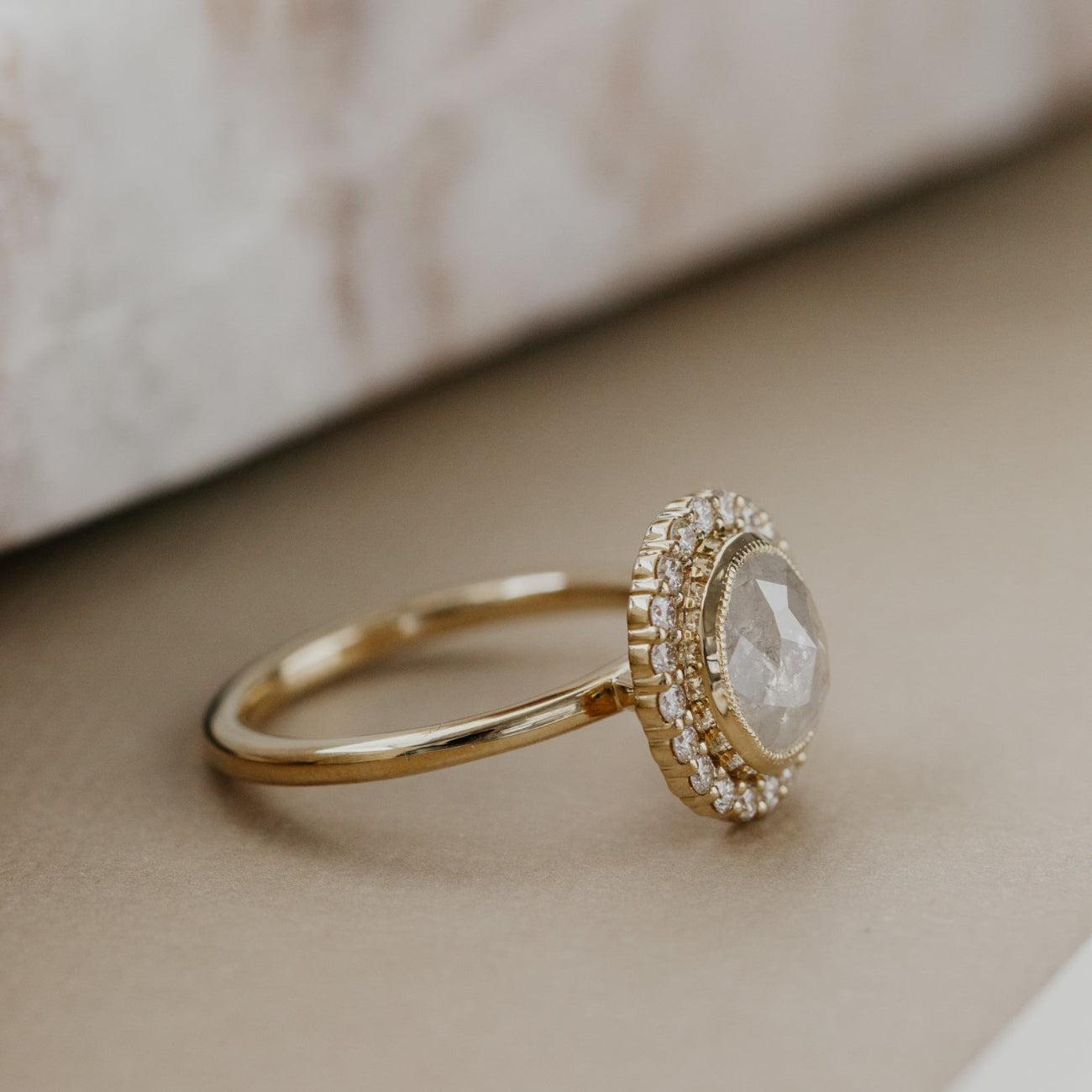 Gold ring with a oval rustic diamond and a diamond halo on a neutral background.