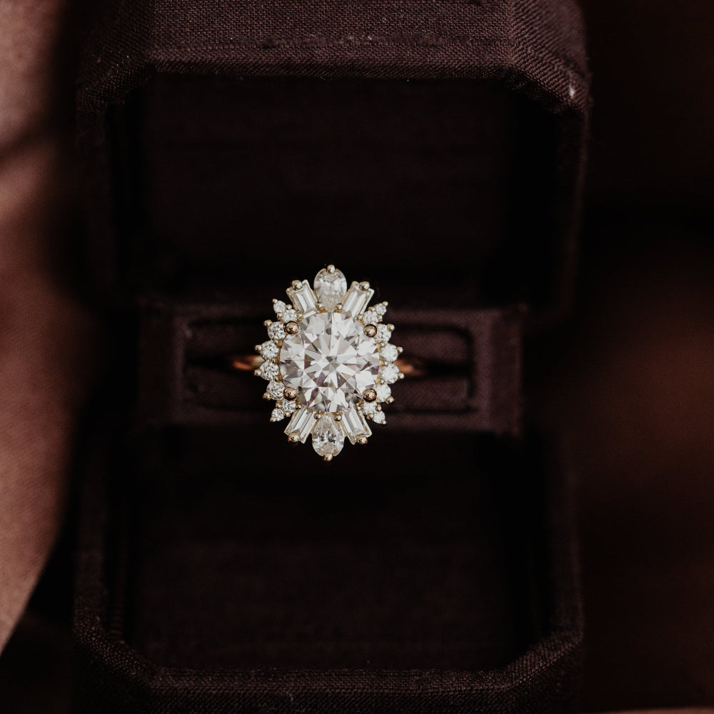 14k gold round Celeste engagement ring with a central diamond surrounded by a starburst halo of pear, baguette and round diamonds, displayed on a black box.