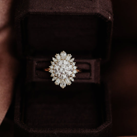 14k gold round Celeste engagement ring with a central diamond surrounded by a starburst halo of pear, baguette and round diamonds, displayed on a black box.