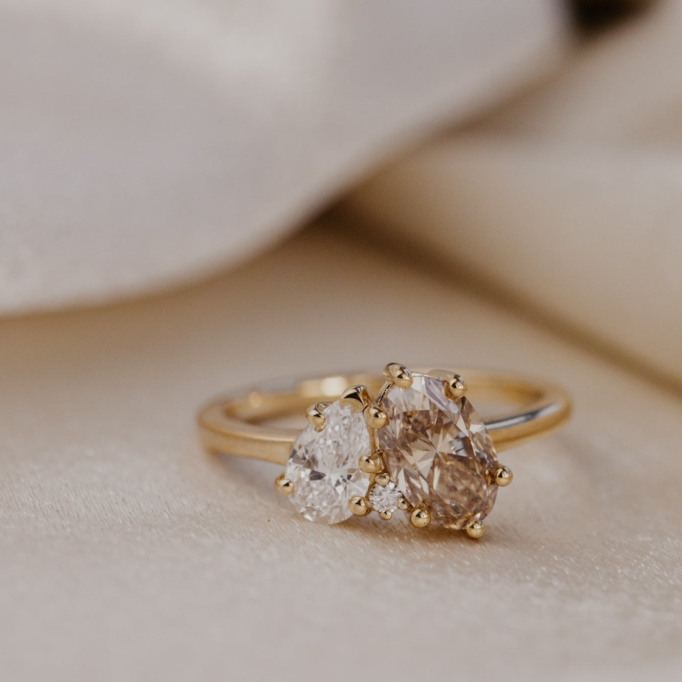 Gold toi et moi ring with a oval champagne diamond and a pear lab grown diamond on a beige surface.