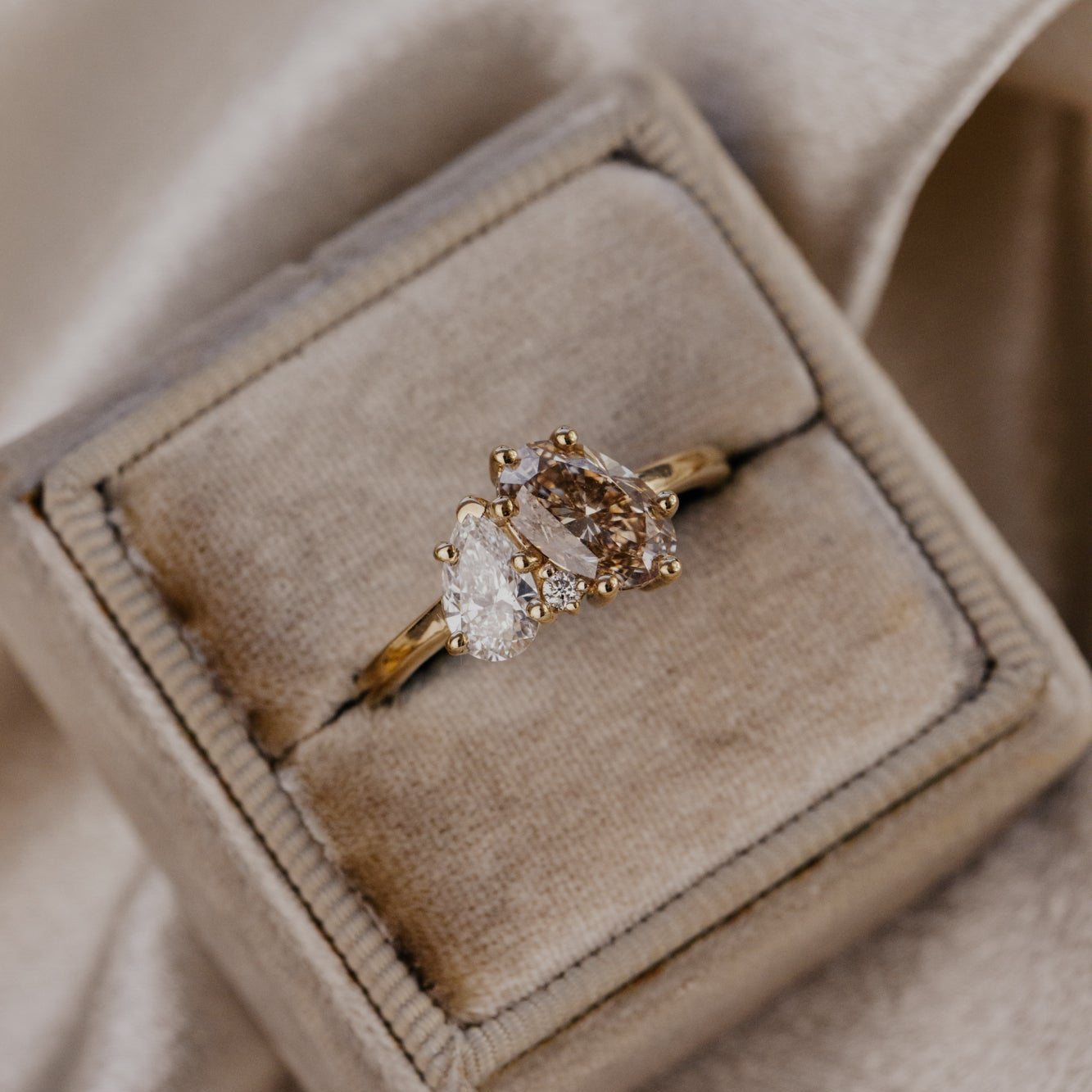 Champagne and lab grown diamond toi et moi ring in a beige velvet box on a neutral background.