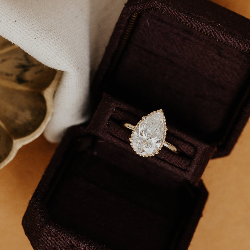 A 14k gold Charlotte pear-shaped diamond ring displayed in a dark velvet box with a beaded texture around the stone, reflecting a sophisticated design.