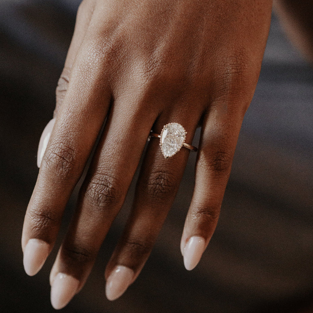 Hand wearing a Charlotte pear-shaped diamond engagement ring with a blurred background