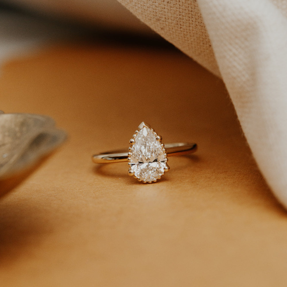 Gold engagement ring with a Charlotte pear-shaped diamond on a beige surface.