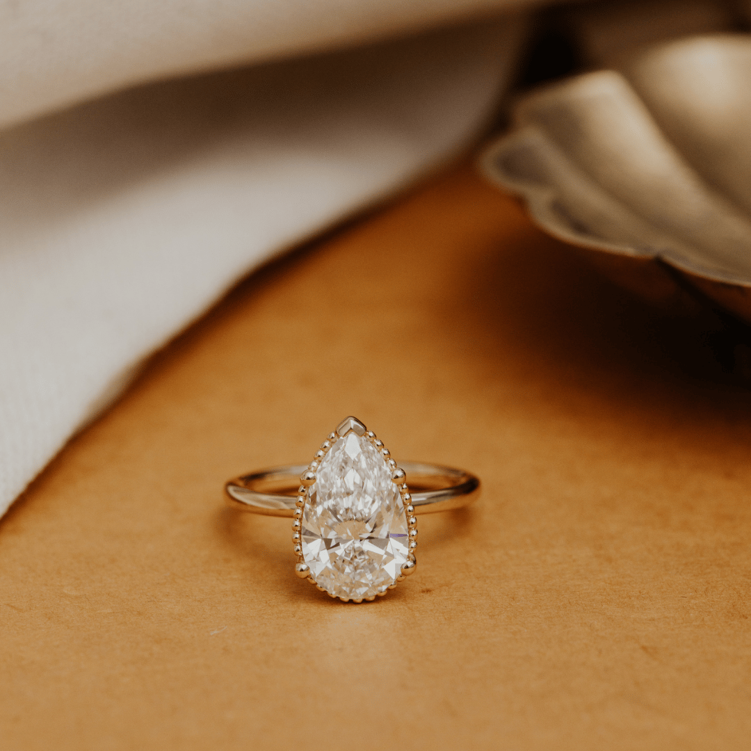 Gold engagement ring with a Charlotte pear-shaped diamond on a beige surface.