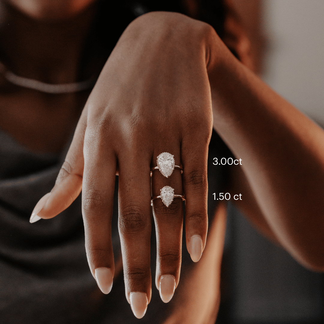 Hand wearing two Charlotte pear-shaped diamond rings with visible carat weights on a blurred background