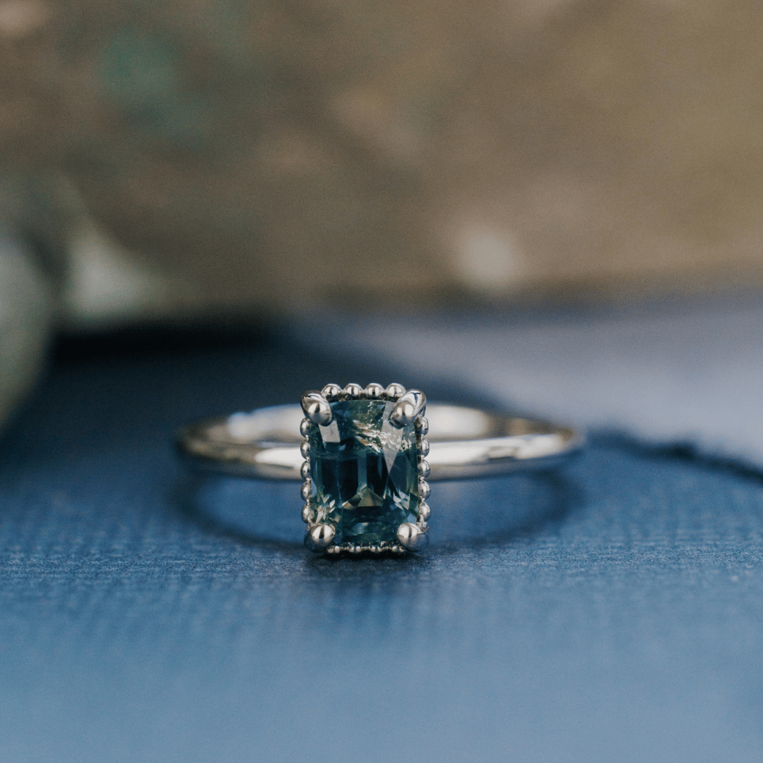 A 14k white gold ring featuring a blue sapphire gemstone, viewed from straight on and displayed on a textured blue surface with a soft-focus background.
