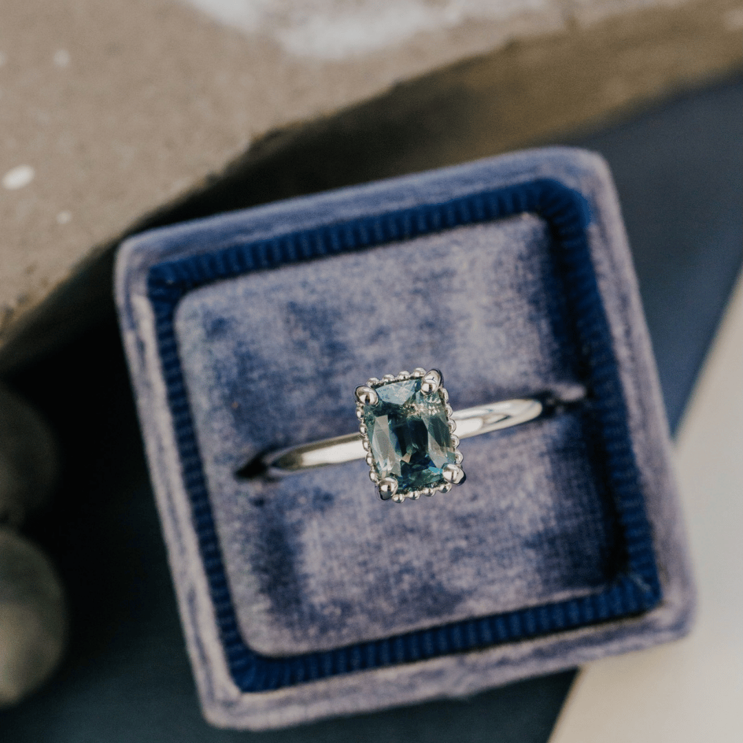 A 14k white gold ring featuring a blue sapphire gemstone, viewed from a ring box and displayed on a textured blue surface with a soft-focus background.