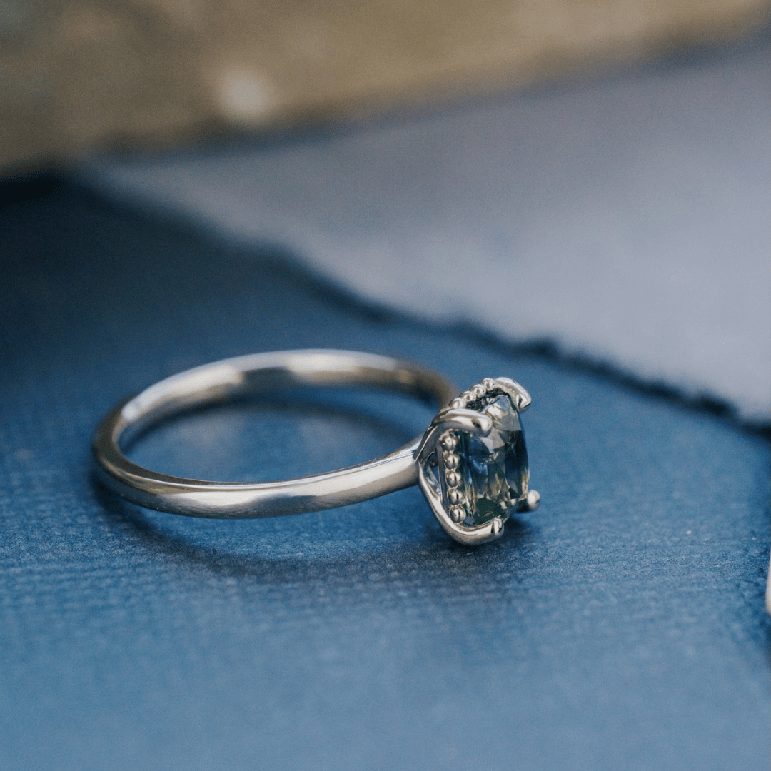 A 14k white gold ring featuring a blue sapphire gemstone, viewed from the side and displayed on a textured blue surface with a soft-focus background.