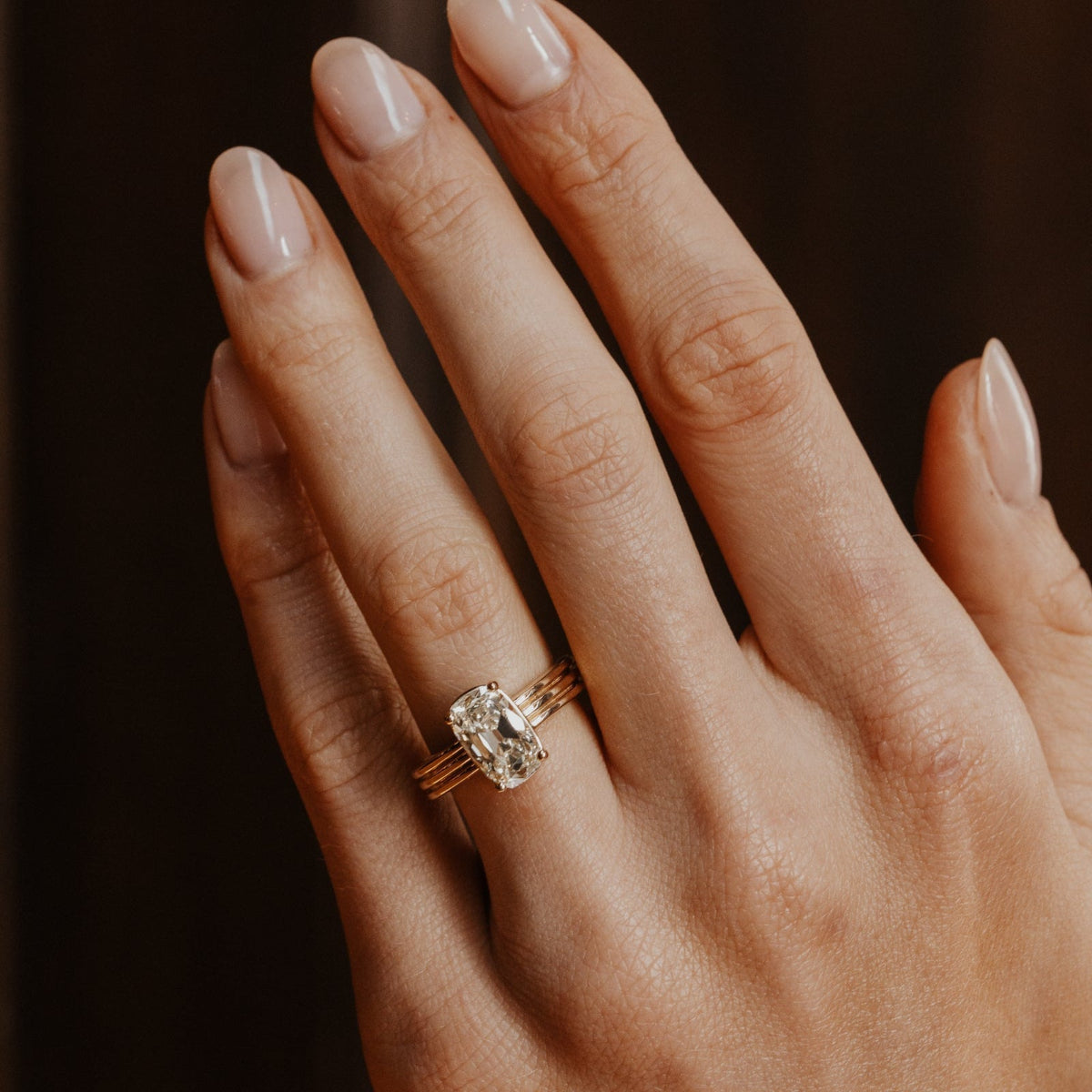 Hand wearing a gold ring with a elongated cushion old mine cut diamond on a dark background.