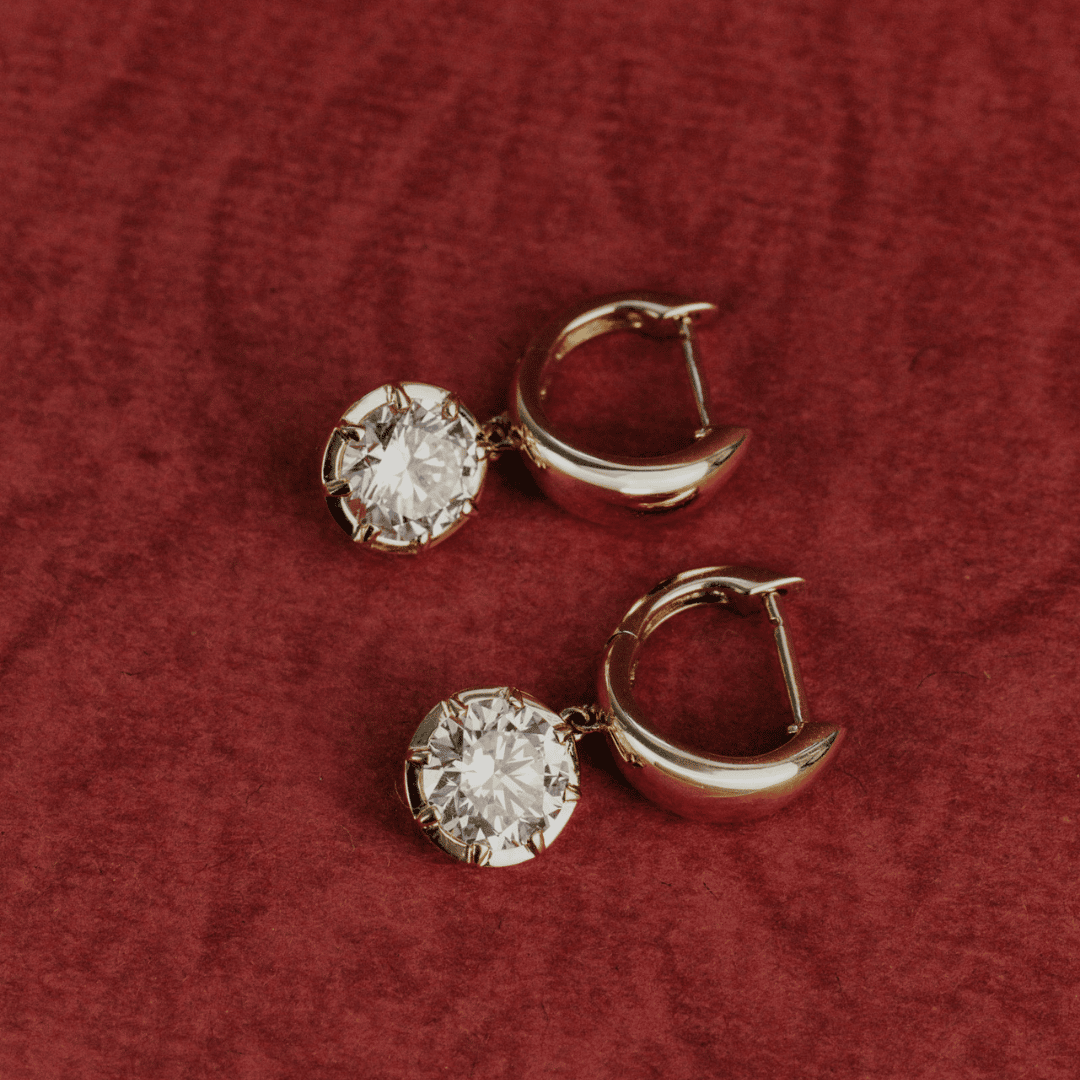 14k gold round cut, lab grown diamond dangle hoop earrings showcased on a textured red surface. 