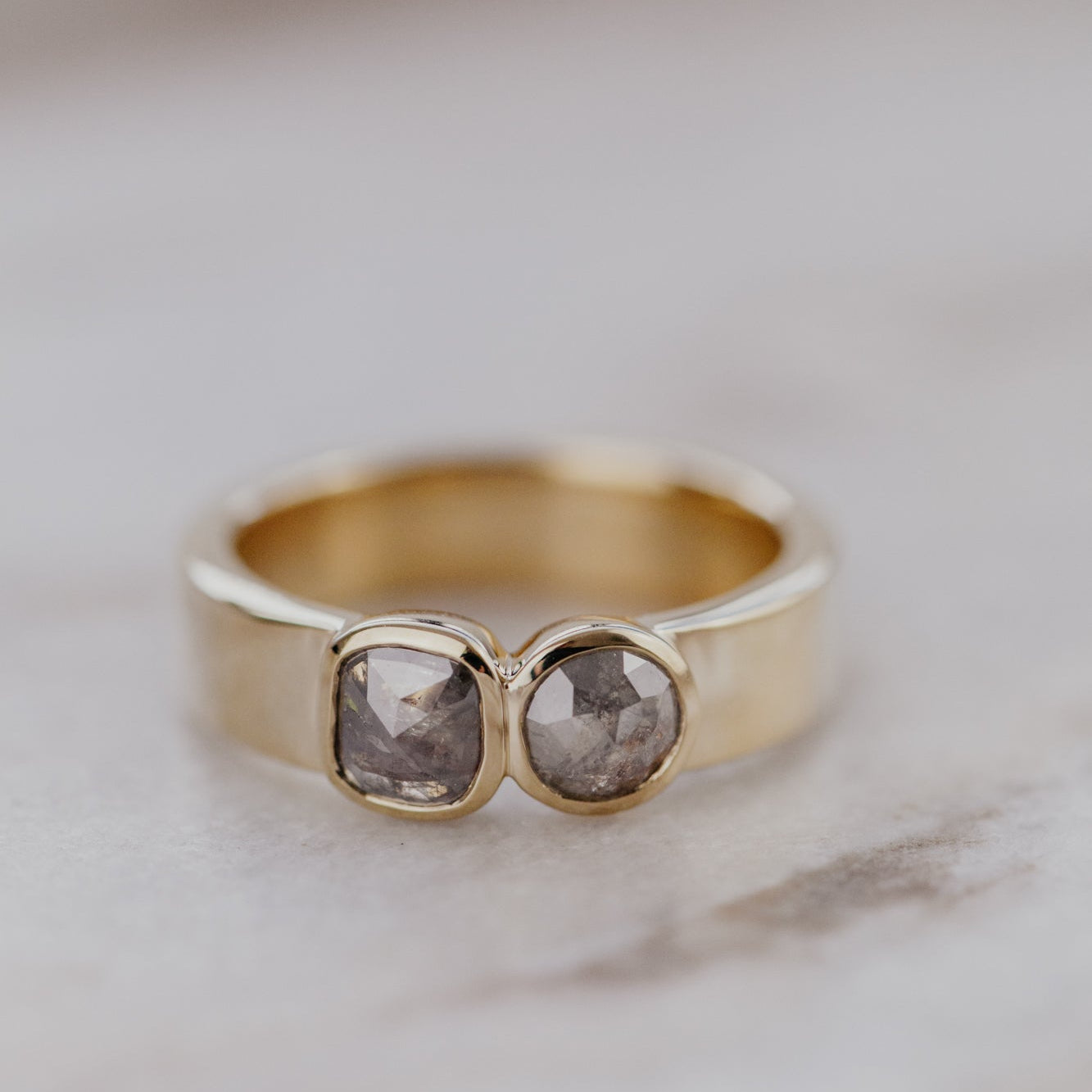 Gold ring with two gray rustic diamond gemstones on a marble surface.