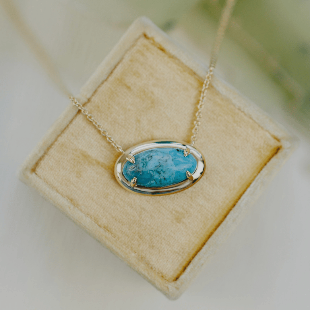 A 14k gold, elongated oval turquoise necklace in a metal border setting on a light colored jewelry box. 