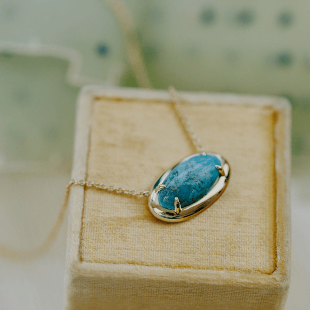 A 14k gold, elongated oval turquoise necklace in a metal border setting on a light colored jewelry box. 
