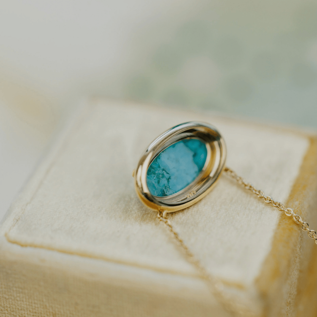 The backside of a 14k gold, elongated oval turquoise necklace in a metal border setting on a light colored jewelry box. 