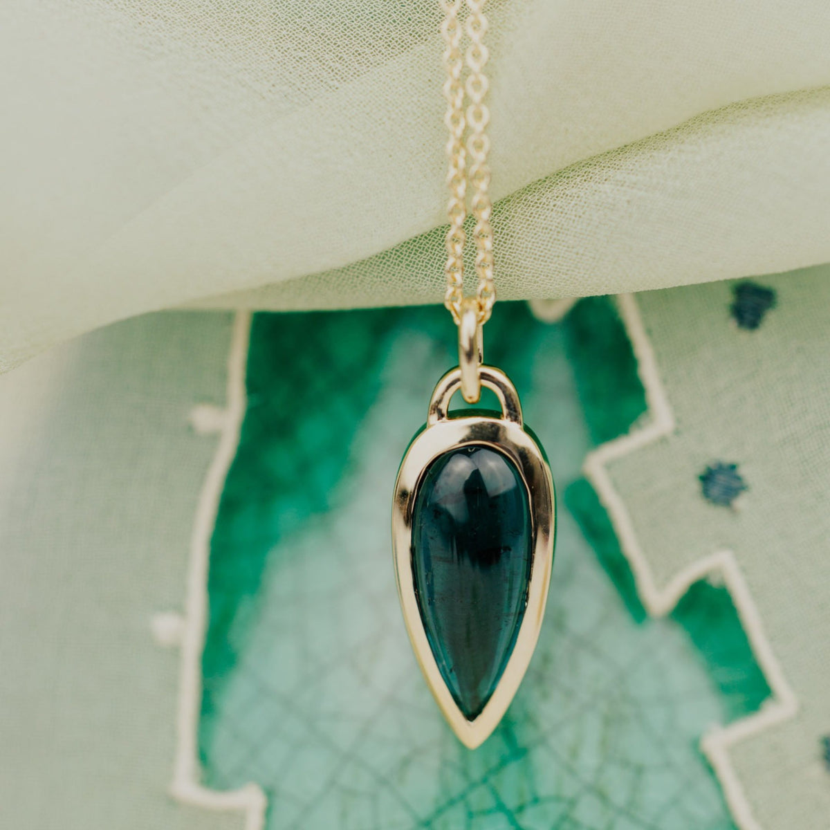 A 14k gold necklace with an elongated pear shaped green tourmaline stone on top of a textured background. 