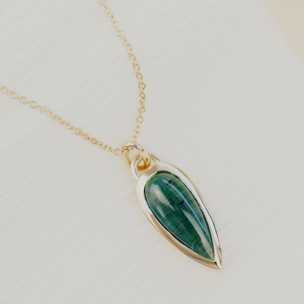 A 14k gold necklace with an elongated pear shaped green tourmaline stone on top of a white background. 