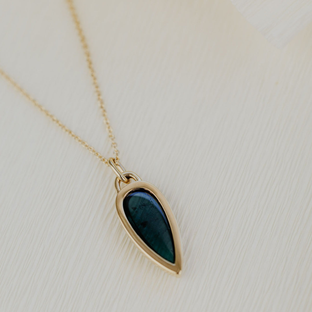 A 14k gold necklace with an elongated pear shaped green tourmaline stone on top of a white background. 