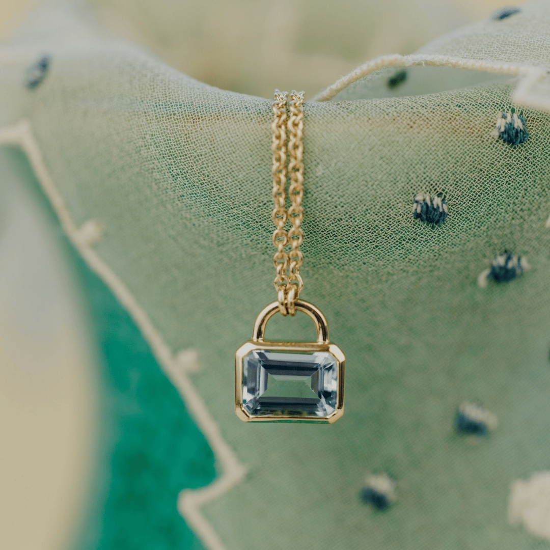 A 14k gold emerald cut bezel set aquamarine lock style necklace set in front of a textured background. 