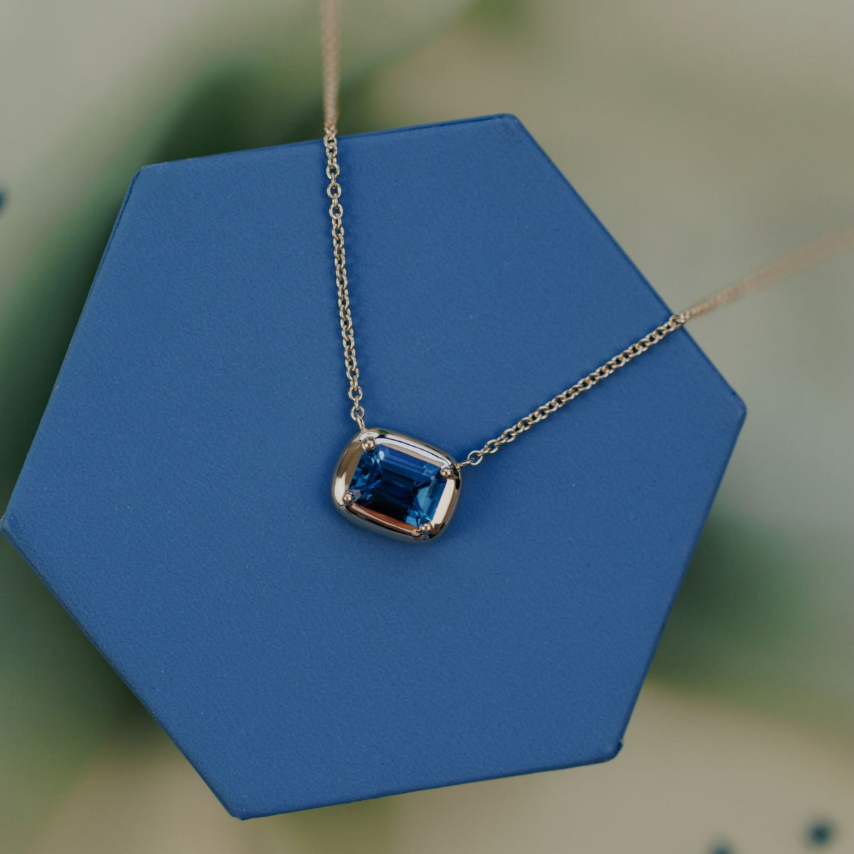 A 14k yellow gold emerald cut, blue topaz necklace on a blue jewelry box with a soft background. 