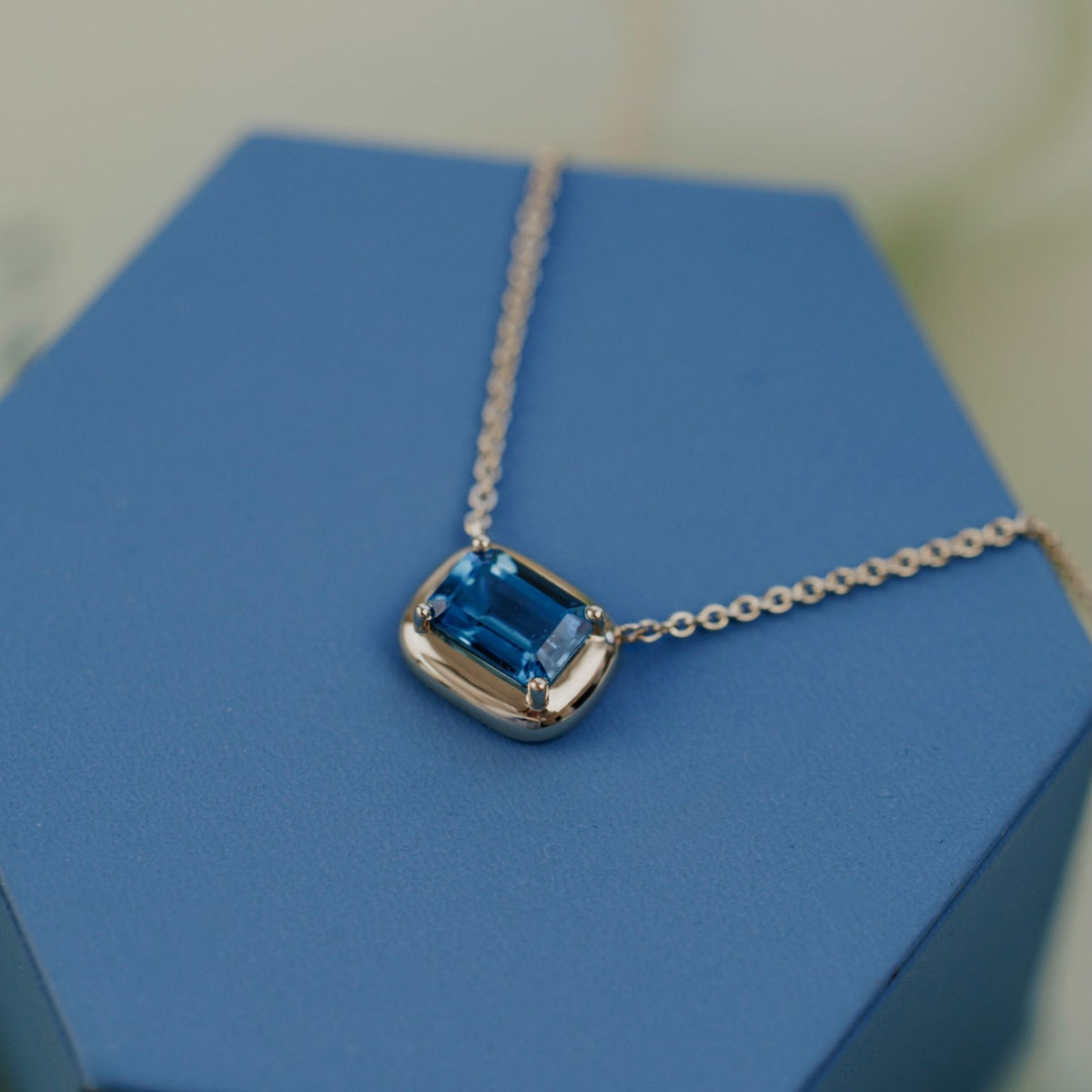 A 14k yellow gold emerald cut, blue topaz necklace on a blue jewelry box with a soft background. 