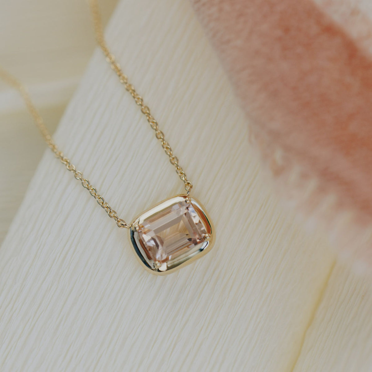An emerald cut morganite necklace in a gold setting set on a soft white background with red fabric adjacent. 