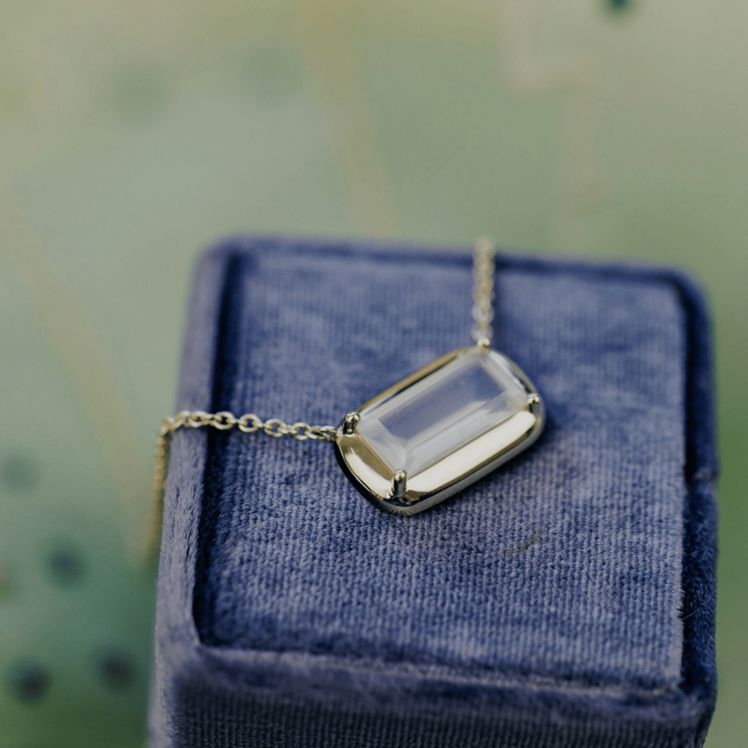 A 14k yellow gold emerald cut moonstone necklace on a soft blue jewelry box on top of a textured background. 