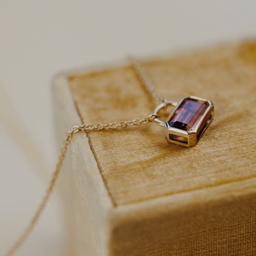 A 14k yellow gold emerald-cut, pink tourmaline stone in a lock style design draped over a soft jewelry box. 