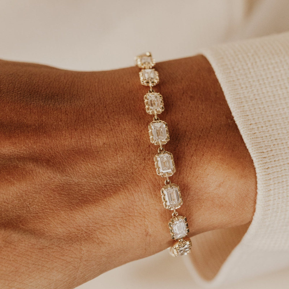 Gold tennis bracelet with emerald cut lab grown diamonds on a wrist against a neutral background.