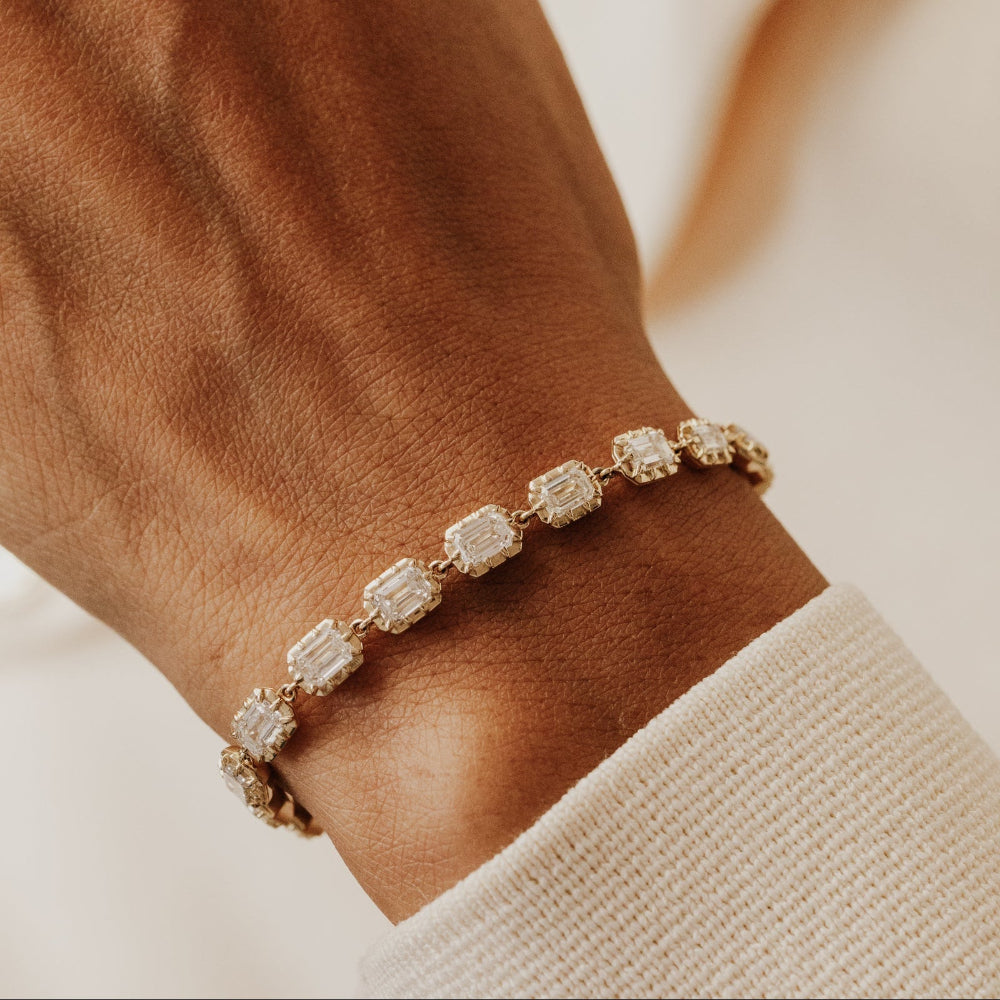 Gold tennis bracelet with emerald cut lab grown diamonds on a wrist against a neutral background.