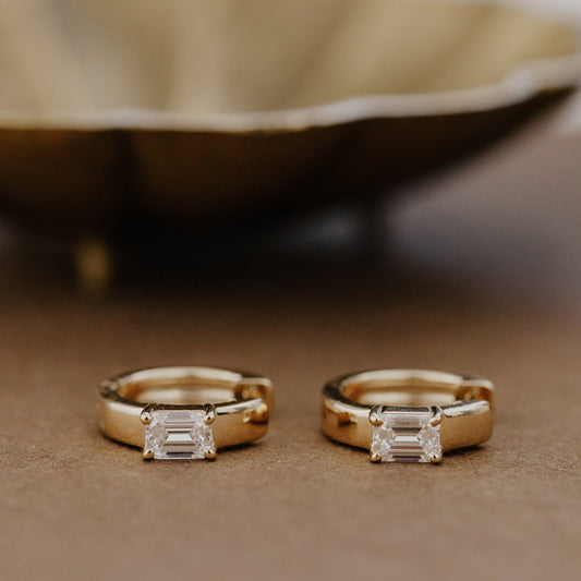 Two gold huggie hoop earrings with emerald cut lab grown diamonds on a brown surface.