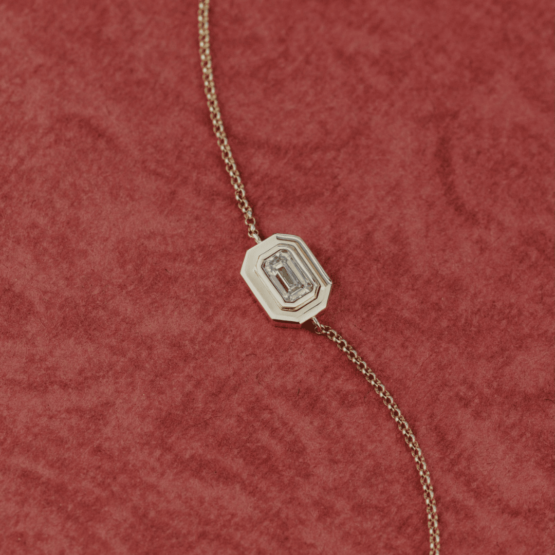 A close up of a 14k gold emerald cut, lab grown diamond chain bracelet showcased on a textured red surface. 