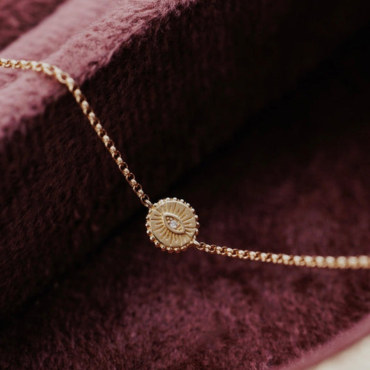 A delicate gold chain bracelet with a round sunburst pendant featuring an eye motif and a small natural diamond, displayed on a textured dark burgundy velvet surface.