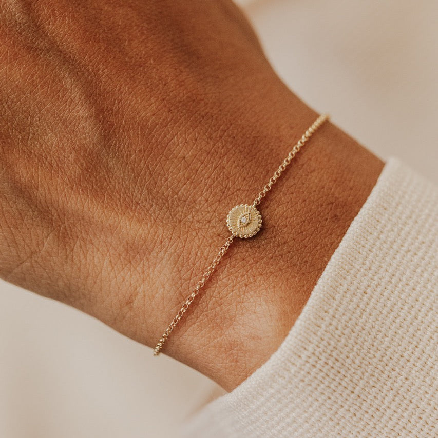 A delicate gold chain bracelet with a small evil eye design with a natural diamond, shown worn on a person's wrist over a white knit cuff.