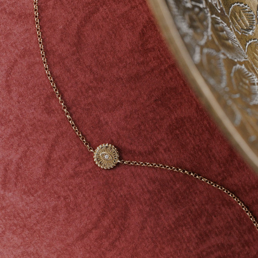 A delicate gold chain bracelet with a small round sunburst pendant featuring a natural diamond, displayed on a textured dark red velvet surface.
