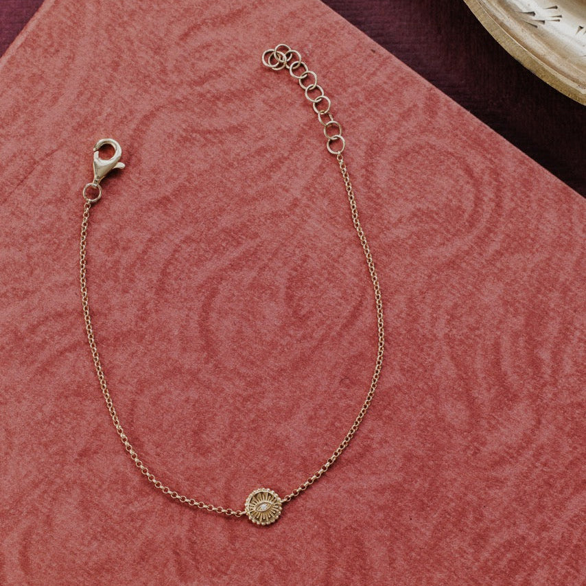 A delicate gold chain bracelet with a small circular sunburst charm featuring a natural diamond, displayed on a textured pink fabric background.