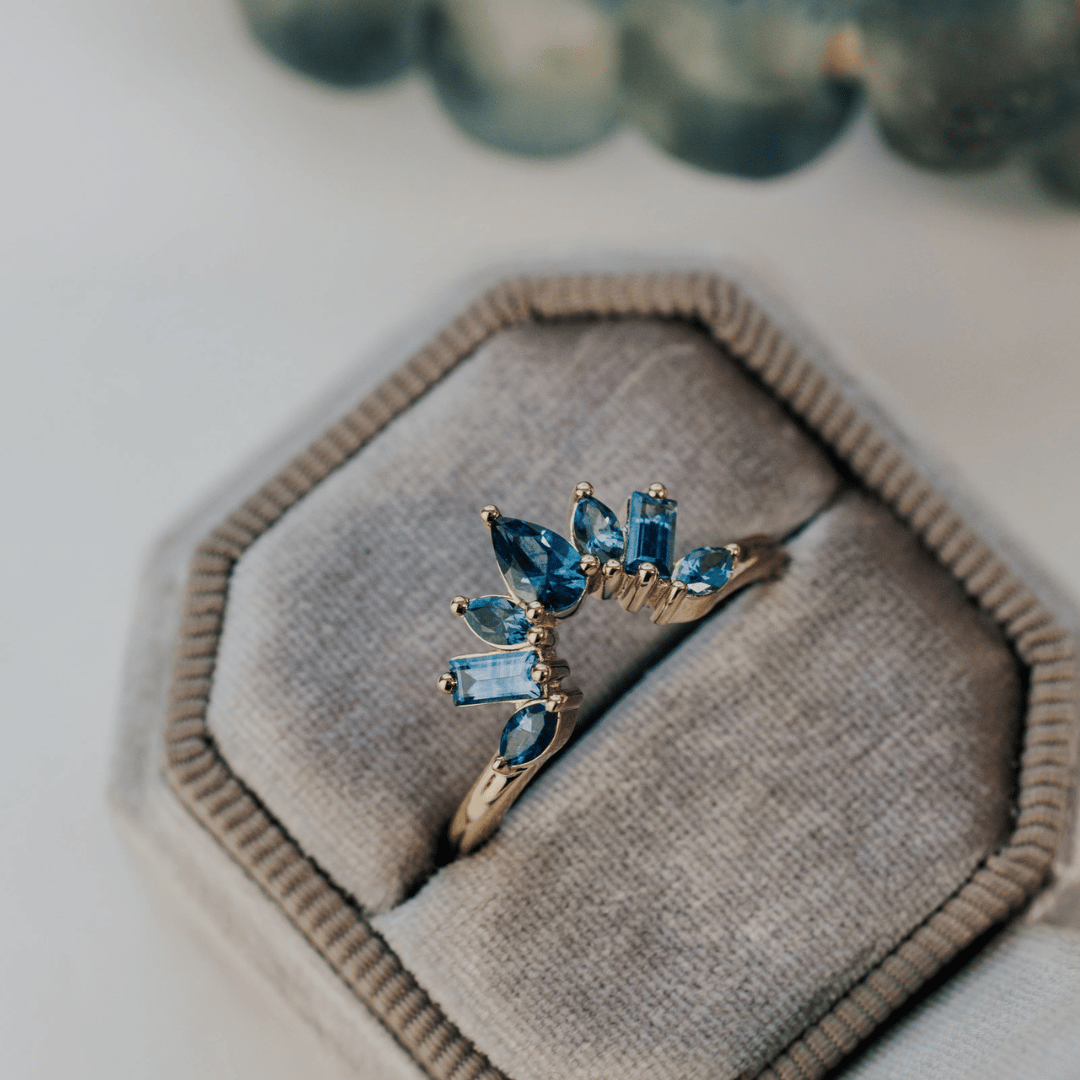 A 14k gold band-style ring with multiple sapphire stones set in a starburst design, shown in a light grey velvet ring box. 