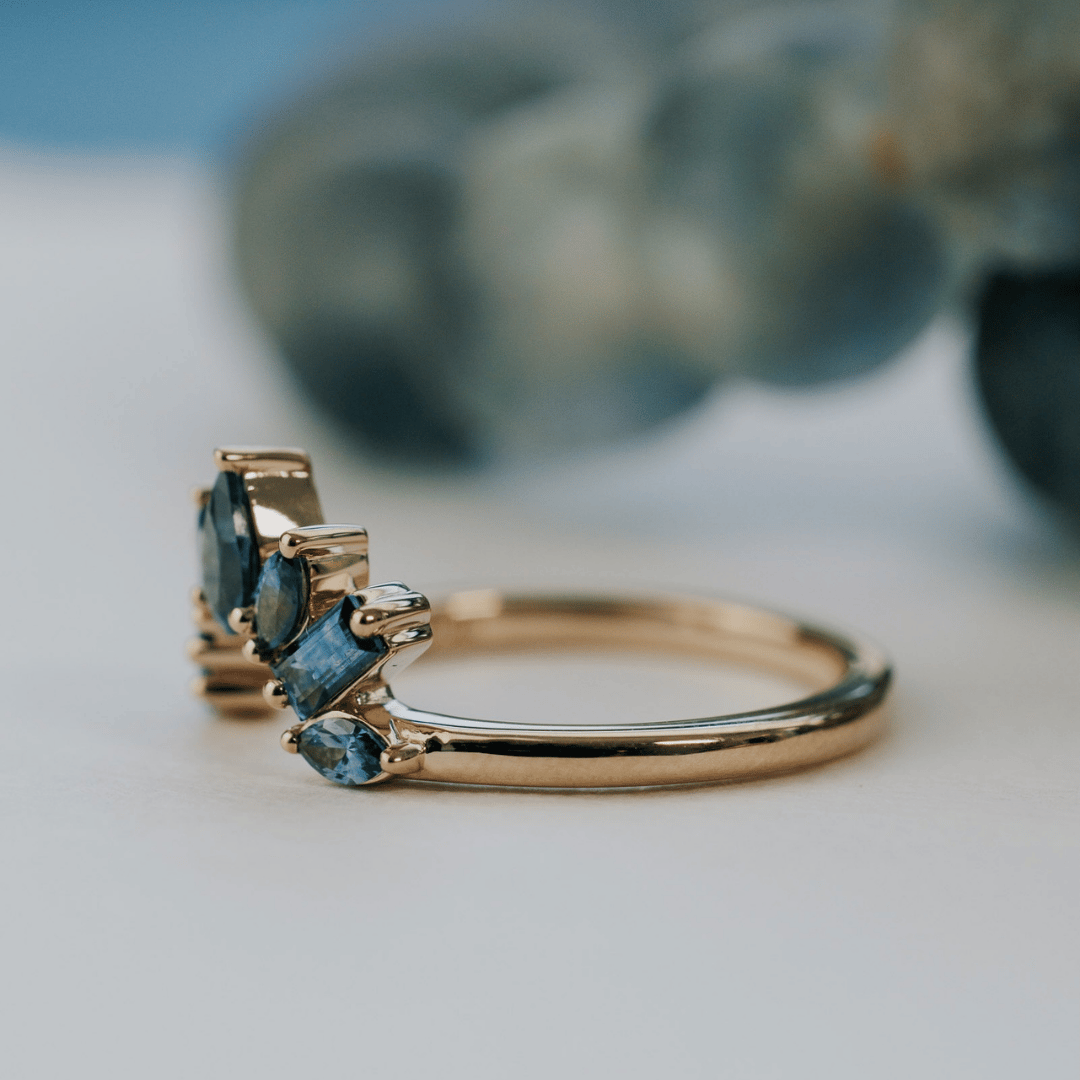 A 14k gold band-style ring with multiple sapphire stones set in a starburst design, angeled to the side, shown on a soft white surface.