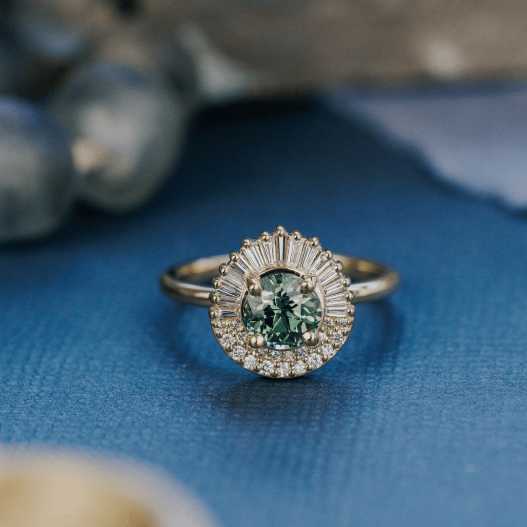 A 14k gold ring featuring a green/blue sapphire gemstone, displayed on a textured blue surface with a soft-focus background.