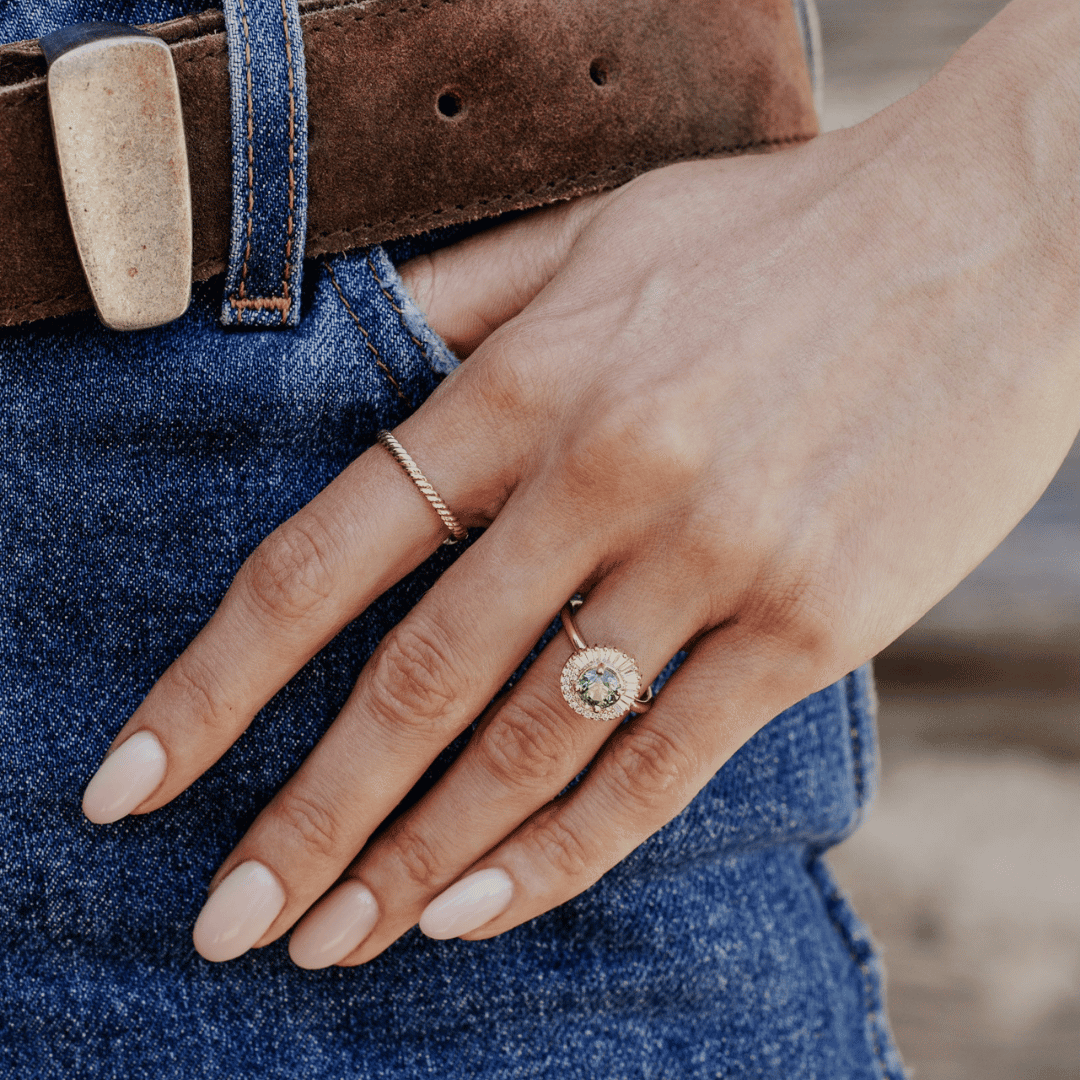 A 14k gold ring featuring a green/blue sapphire gemstone, displayed on the ring finger of a hand model.