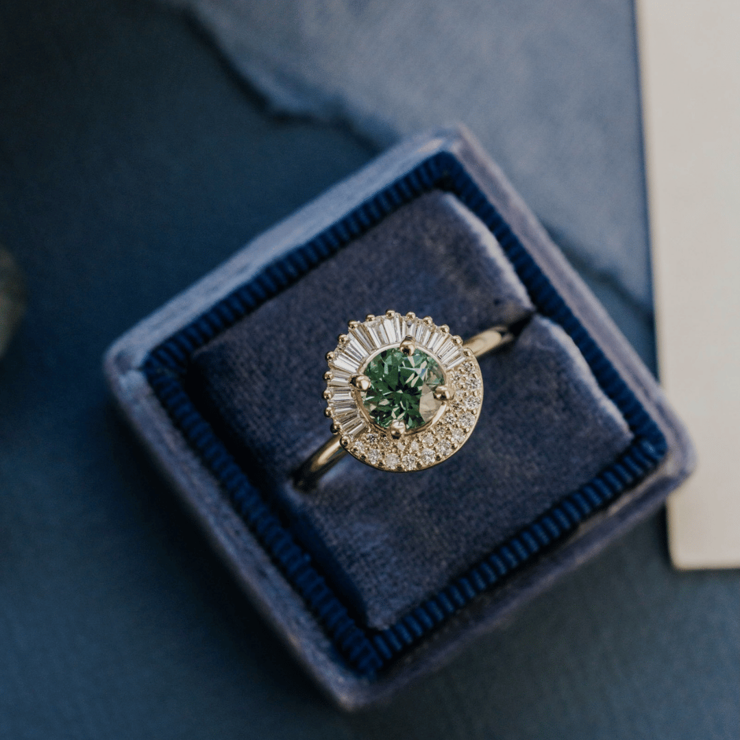 A 14k gold ring featuring a green/blue sapphire gemstone, displayed in a ring box on a textured blue surface with a soft-focus background.