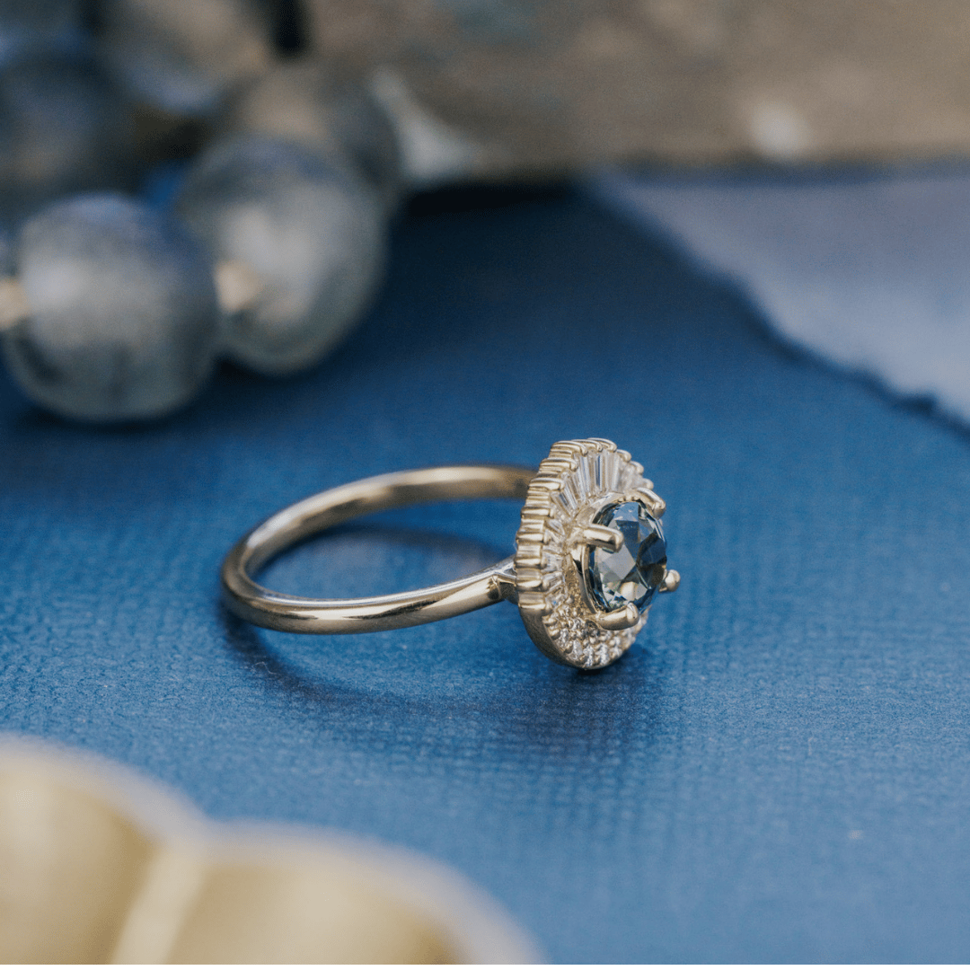 A 14k gold ring featuring a green/blue sapphire gemstone from a side view, displayed on a textured blue surface with a soft-focus background.
