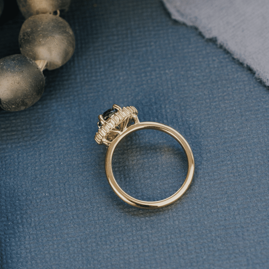 A 14k gold ring featuring a green/blue sapphire gemstone from a top view, displayed on a textured blue surface with a soft-focus background.