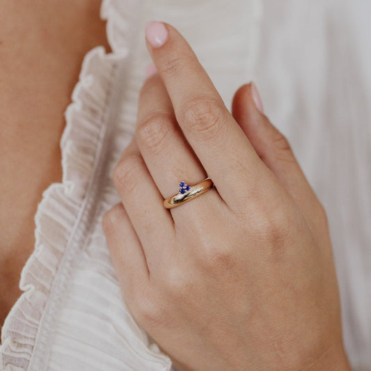 Hand wearing a gold ring with a small blue gemstone, against a blurred white fabric background.